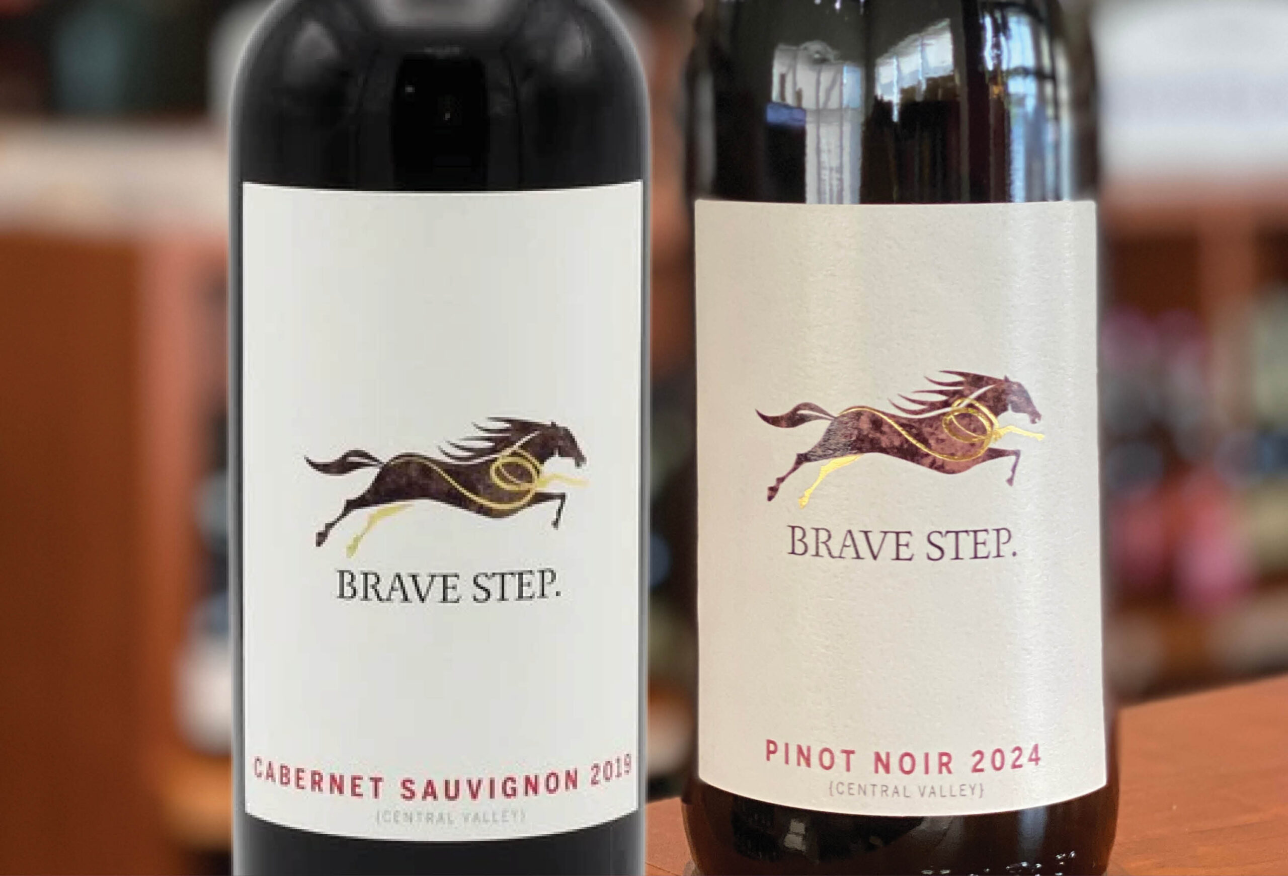 Two wine bottles labeled Brave Step. The left is a Cabernet Sauvignon 2019, the right is a Pinot Noir 2024. Both feature a running horse logo, with shelves and bottles blurred in the background.