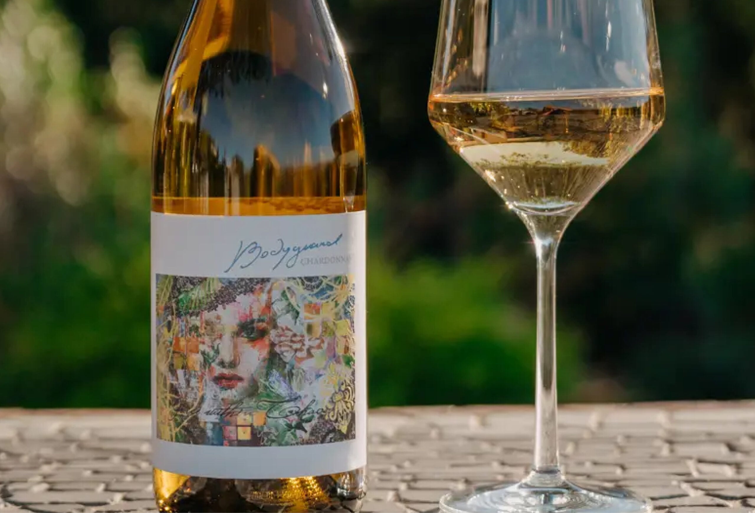 A bottle of white wine with a colorful, artistic label next to a filled wine glass sits on a sunlit outdoor table with a blurred green background—perfect for savoring our Buy One Get One Free Wines offer.