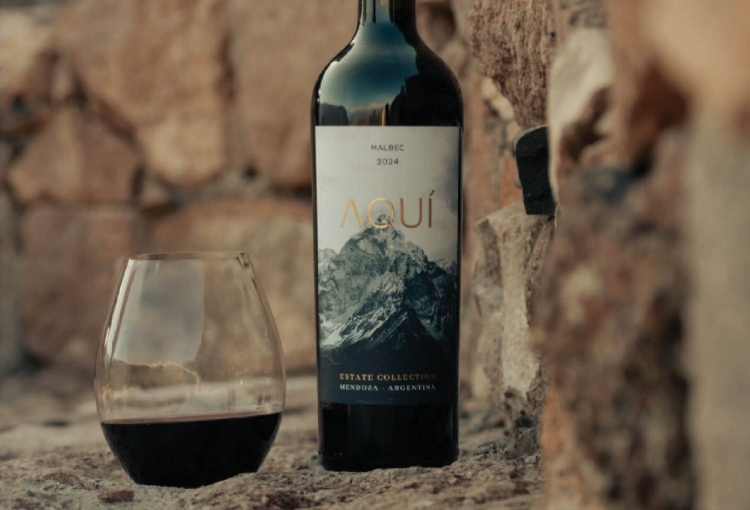 A bottle of Aquí Malbec 2024 wine and a glass of red wine rest on a stone surface, with a rustic stone wall in the background. The wine label features a mountain image and text indicating it is from Mendoza, Argentina.