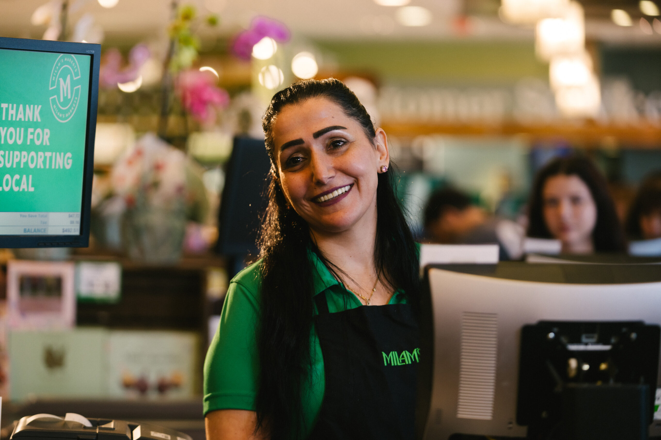 A smiling woman in a green shirt and black apron stands behind a store counter, with a sign nearby that reads, Thank you for supporting local. Blurred people and flowers are visible in the background.