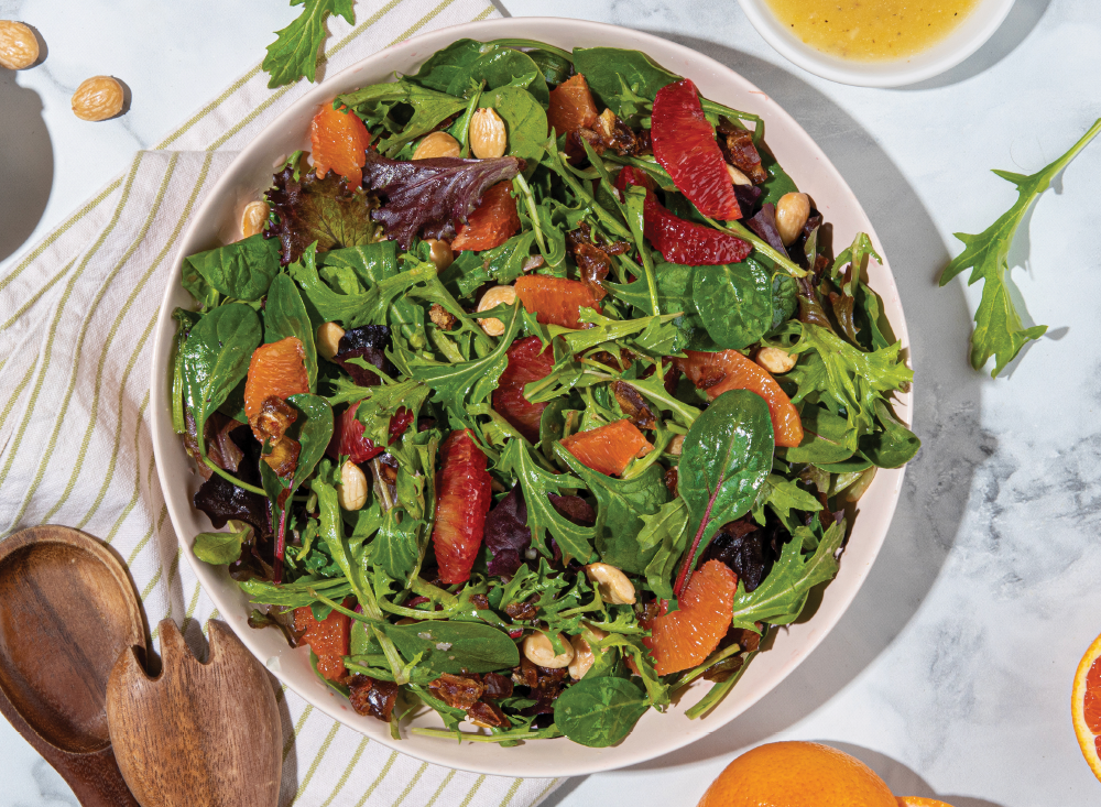 A bowl of mixed organic greens salad with arugula, citrus segments, nuts, and dressing sits on a white surface beside wooden salad utensils, a striped napkin, and a small bowl of vinaigrette.